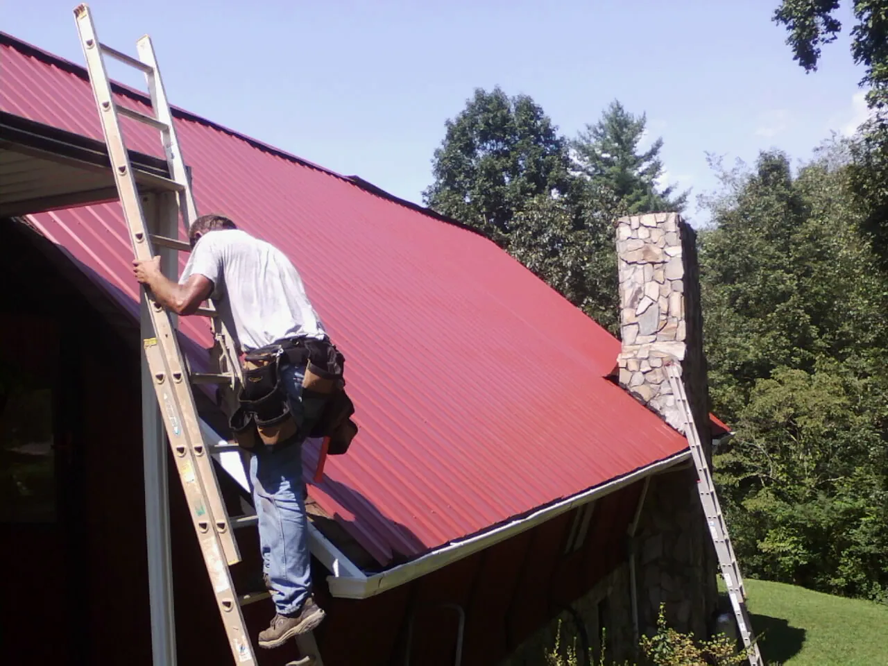 Roofer on a ladder beside a red metal roof and stone chimney — Soffit & Fascia Repair by Roofing Brothers in Torrey